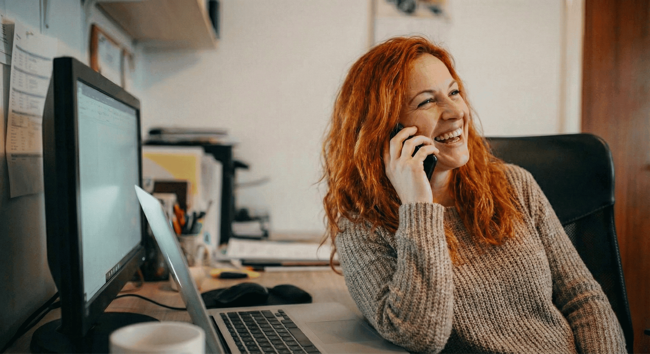 Red head woman on the phone with an employment lawyer in her home office, seemingly happy and smiling. 