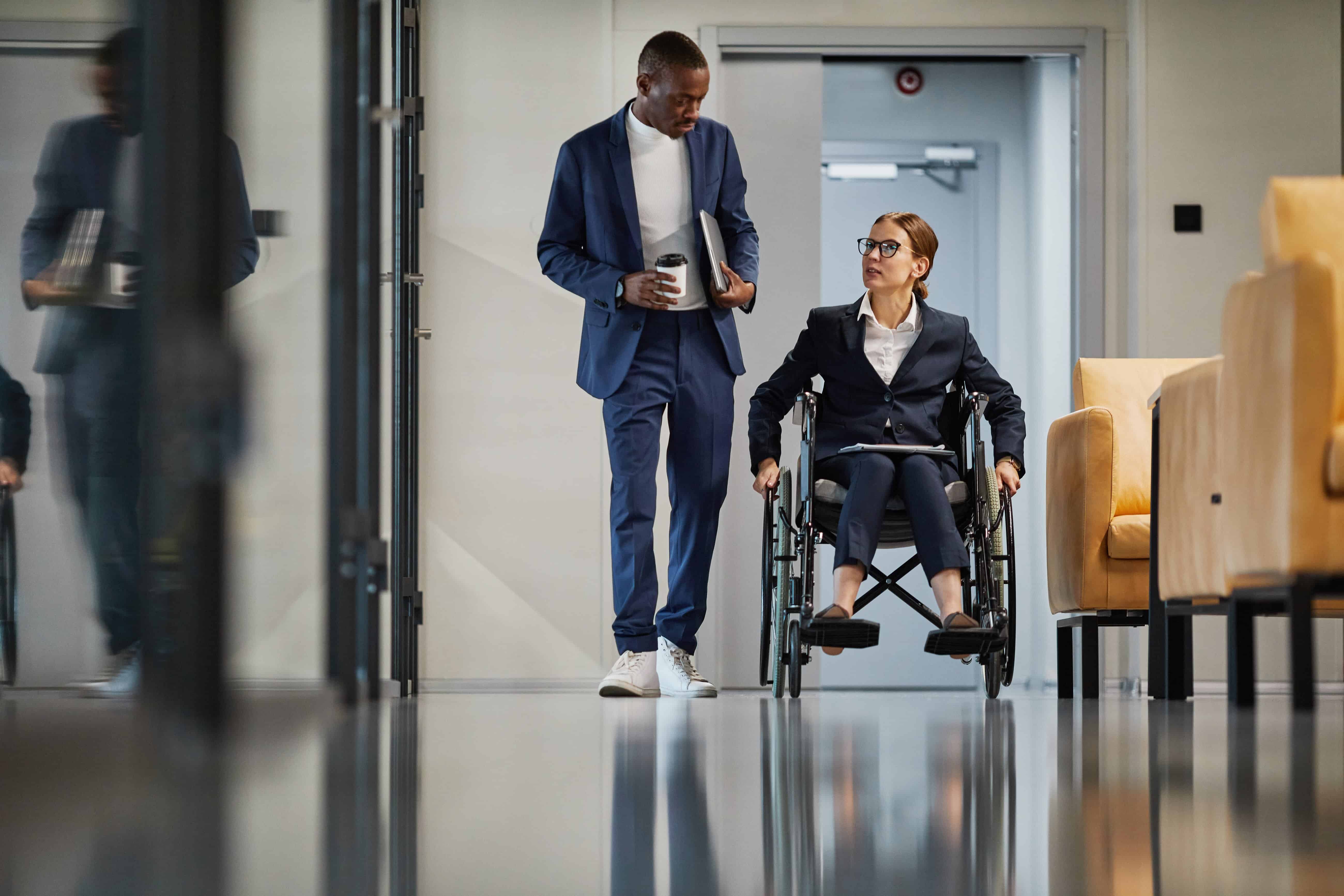 Disabled California female with red hair and glasses in a wheelchair alongside her Black male co-worker in an office hallway